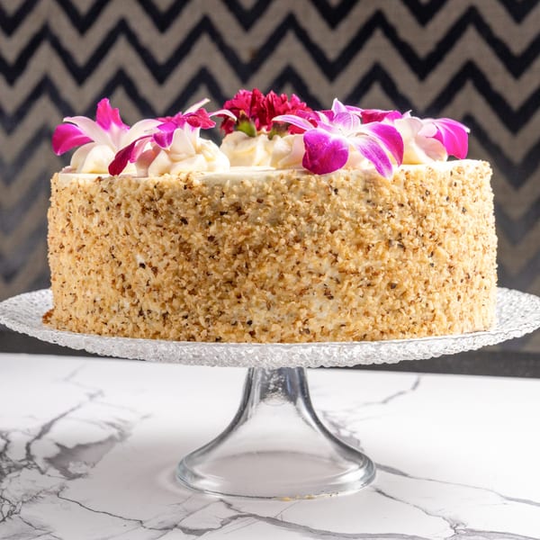 A round cake with nut-covered sides and white frosting topped with pink and purple flowers.
