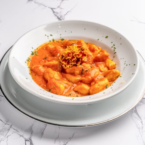 Gnocchi in a vibrant orange sauce, garnished with parsley and an edible flower, on a white marble surface.