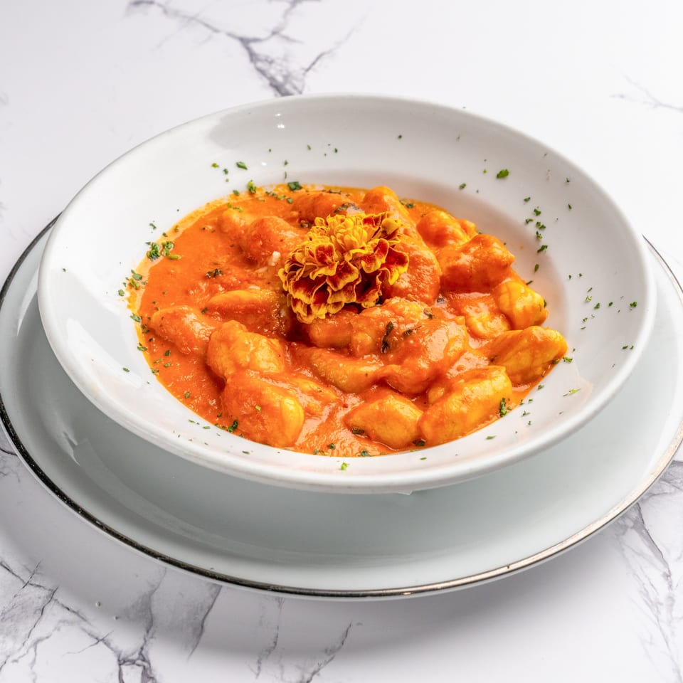 Gnocchi in a vibrant orange sauce, garnished with parsley and an edible flower, on a white marble surface.