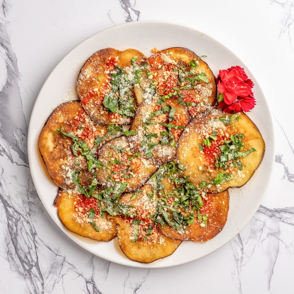 Fried eggplant slices topped with red sauce, grated cheese, and fresh basil, garnished with a red flower.