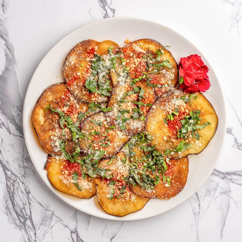 Fried eggplant slices topped with red sauce, grated cheese, and fresh basil, garnished with a red flower.