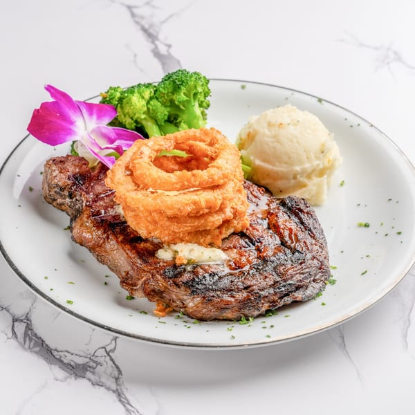 Grilled steak topped with onion rings, served with mashed potatoes, broccoli, and a purple orchid.