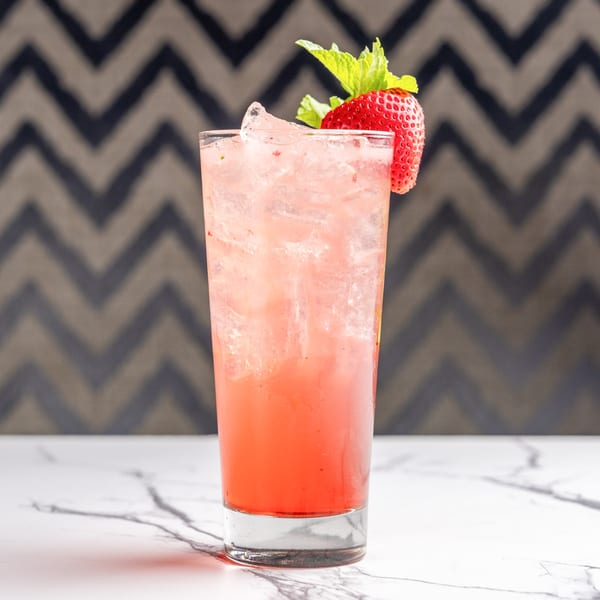 A tall glass of pink strawberry drink with ice, garnished with a fresh strawberry and mint.