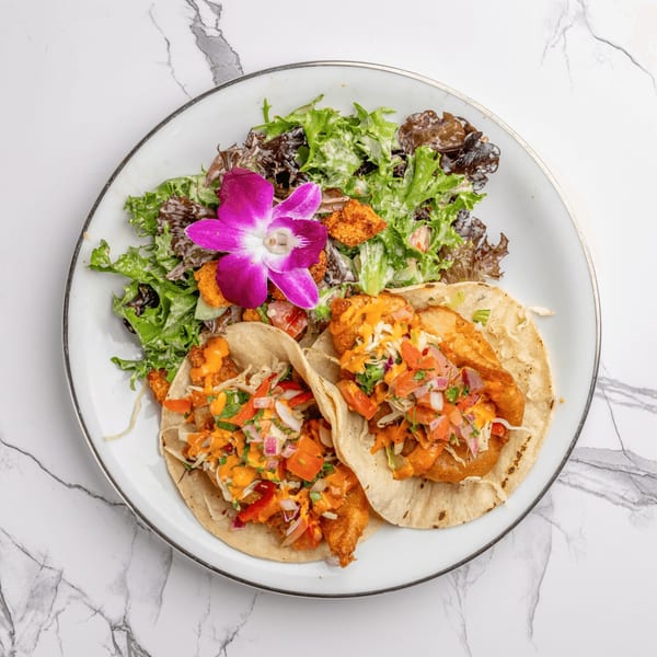 Two fish tacos with salsa and a side salad garnished with a purple orchid on a white plate.