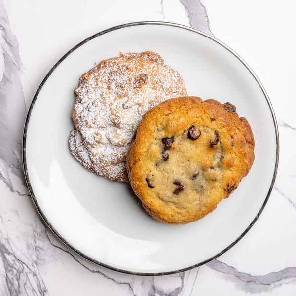Two cookies on a white plate, one chocolate chip and one powdered sugar, on a marble surface.