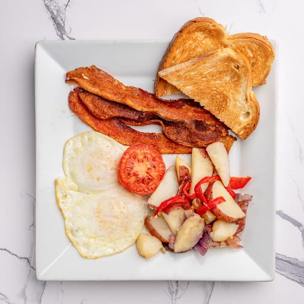 Breakfast plate with crispy bacon, fried eggs, roasted potatoes, grilled tomato, and toast.