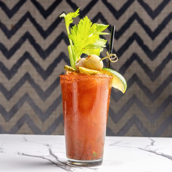 Bloody Mary cocktail garnished with celery, olives, pickles, and a lime wedge on a marble surface.