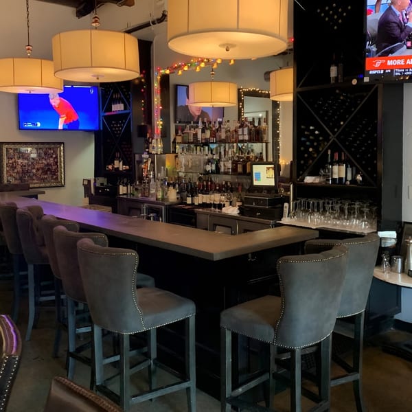 The Bistro K's dimly lit bar area with bottles, glasses, bar stools, and two TVs.