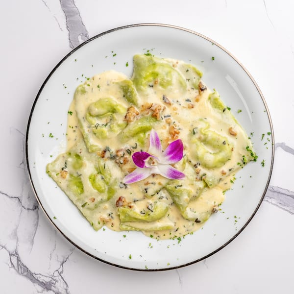 Green ravioli in a creamy sauce with walnuts and a purple orchid garnish on a white plate.