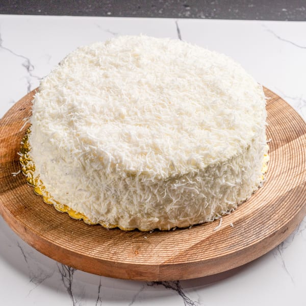 A round white coconut cake on a wooden serving board, against a marble background.