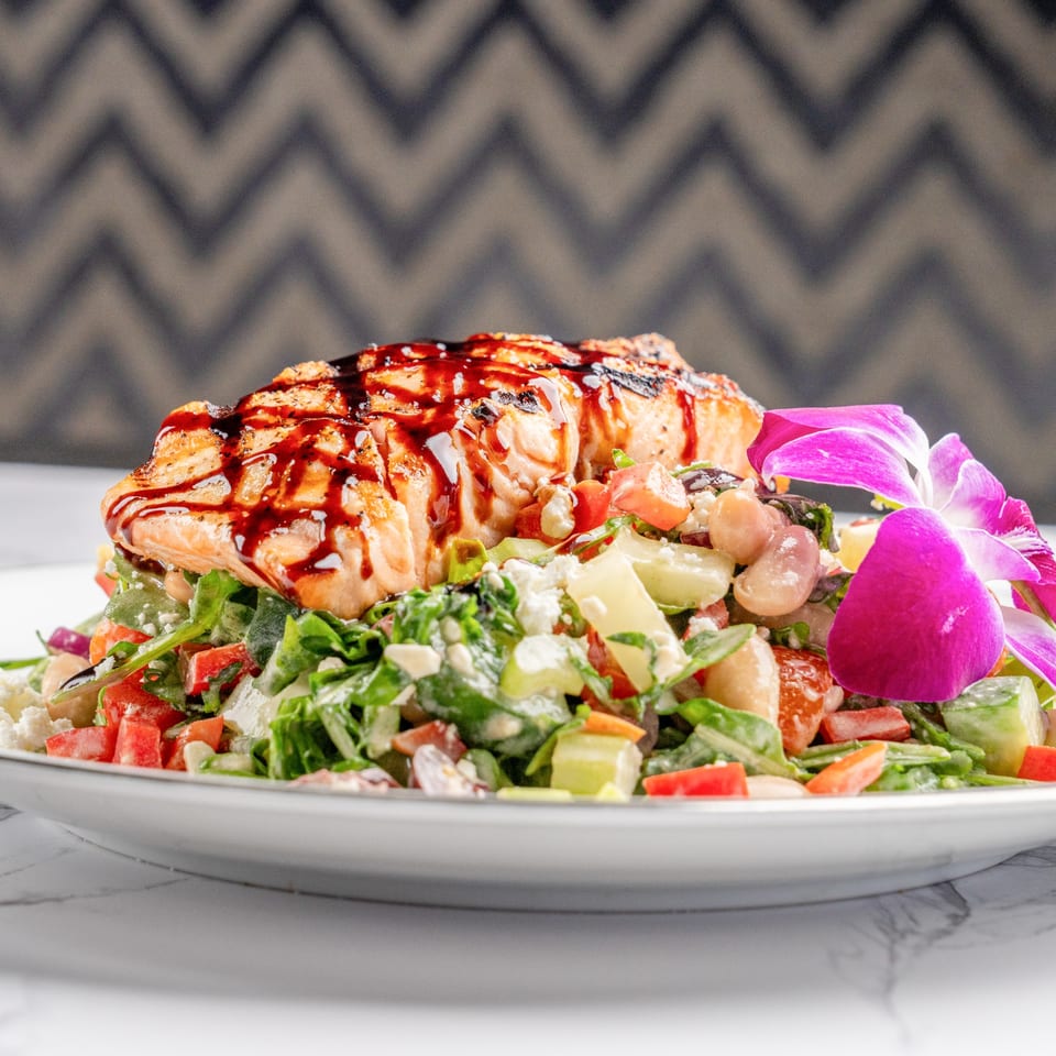 Grilled salmon with balsamic glaze on a fresh salad with feta, beans, and a purple orchid.