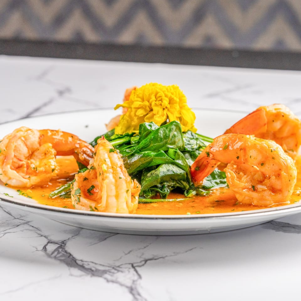 Shrimp scampi with sautéed spinach and a yellow flower garnish on a white plate.
