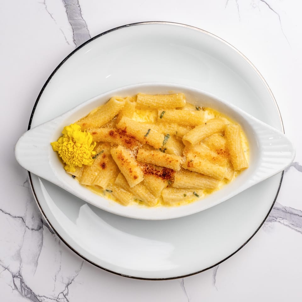 Baked rigatoni pasta with a creamy sauce, topped with breadcrumbs and a yellow flower garnish.
