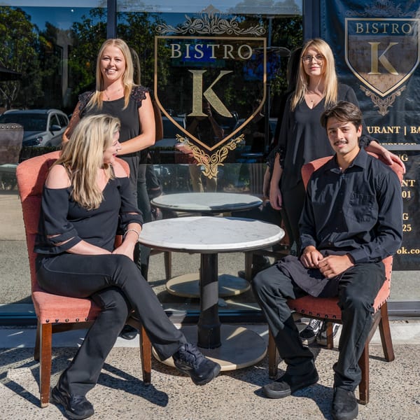 Four Bistro K staff members, two seated and two standing, pose in front of the restaurant's entrance.