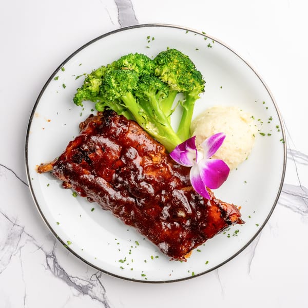 Glazed ribs with broccoli, mashed potatoes, and an orchid garnish on a white plate.