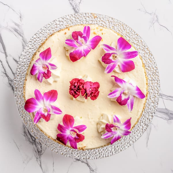 Overhead view of a cheesecake on a glass stand, garnished with pink orchids and red carnations.