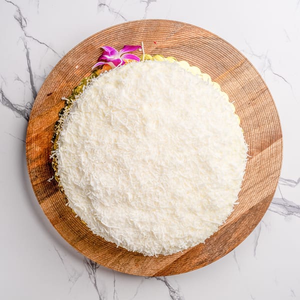 A round coconut cake with white shredded coconut topping and a purple orchid on a wooden board.