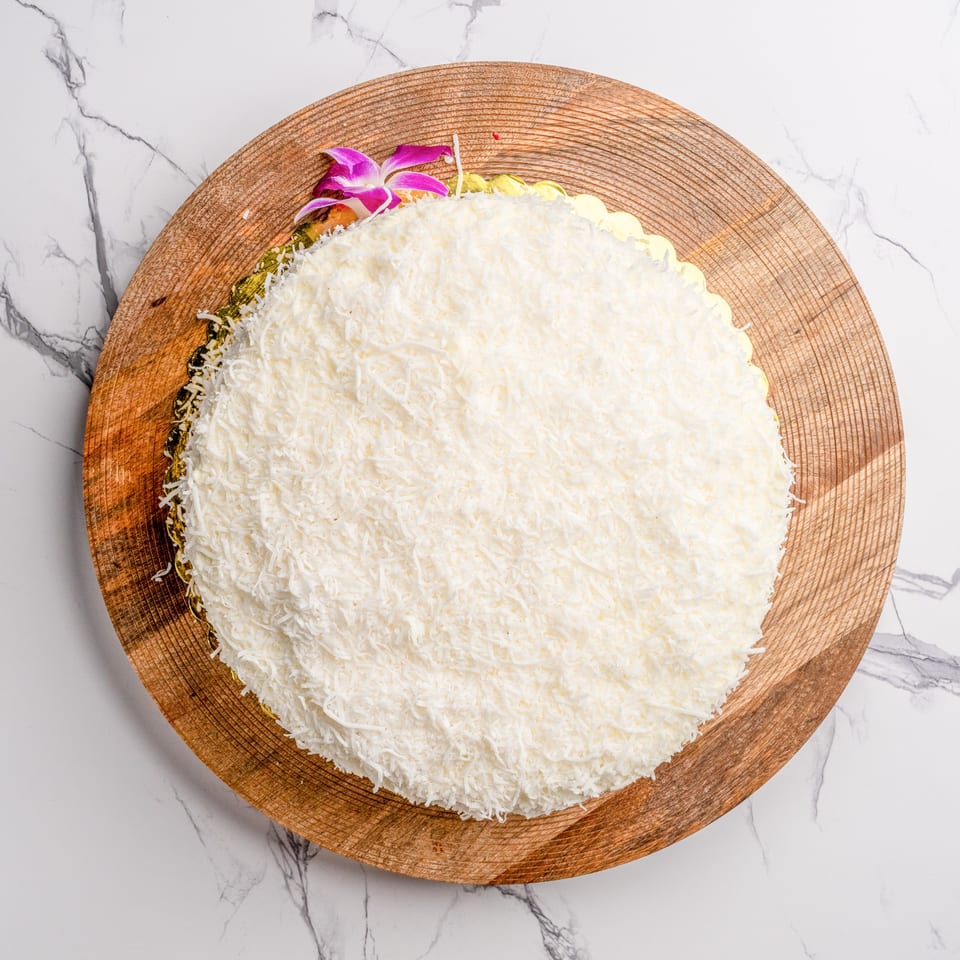 A round coconut cake with white shredded coconut topping and a purple orchid on a wooden board.