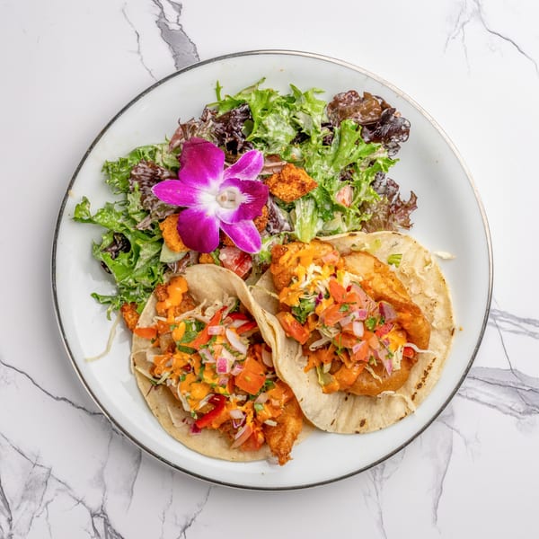 Two fish tacos with salsa and sauce, served with a side salad and purple orchid on a white plate.
