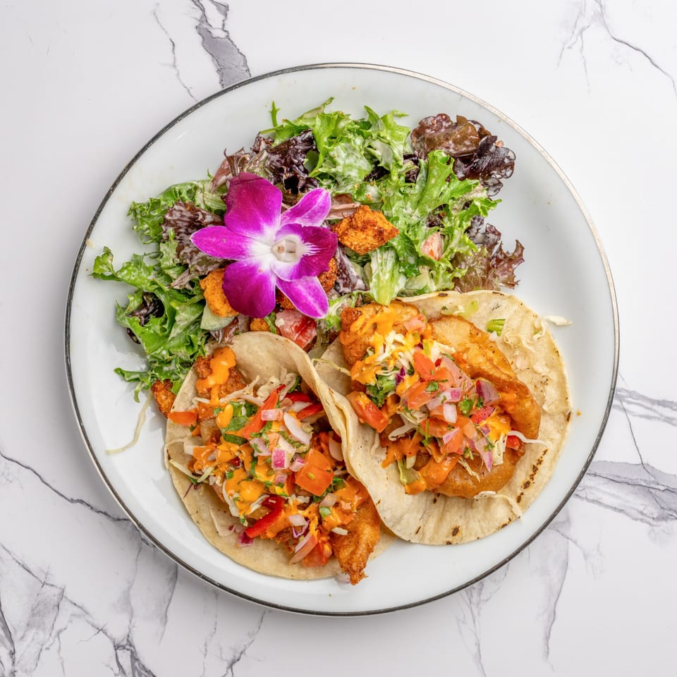 Two fish tacos with salsa and sauce, served with a side salad and purple orchid on a white plate.