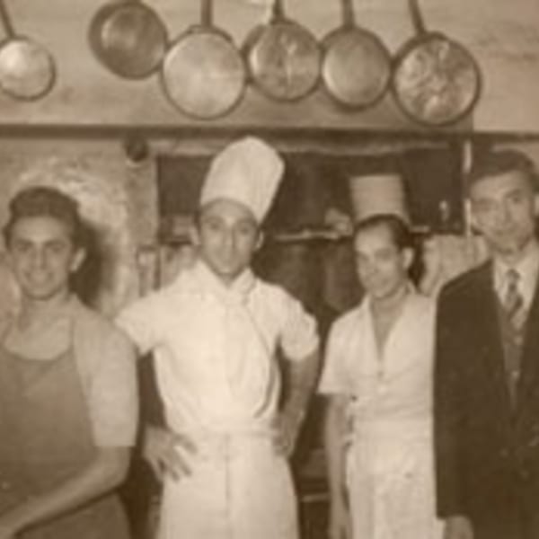 Four men, two in chef's whites, stand in a vintage kitchen with hanging pots.