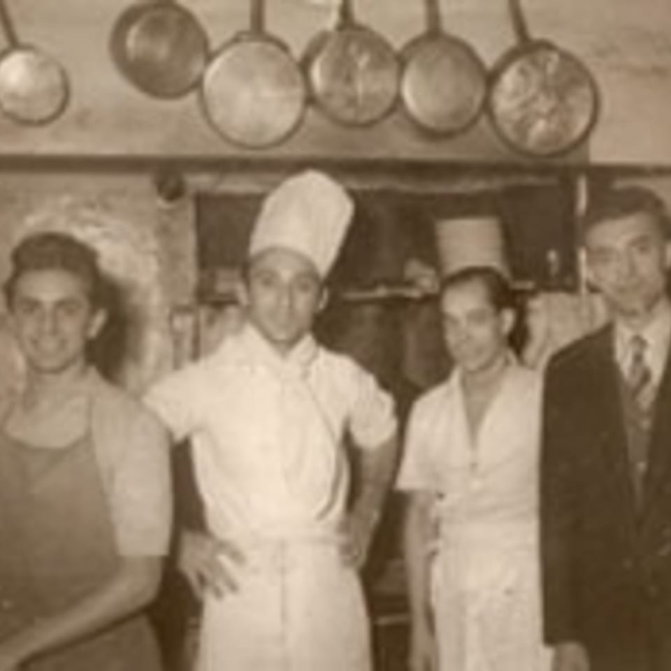 Four men, two in chef's whites, stand in a vintage kitchen with hanging pots.