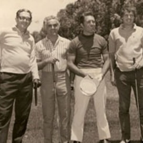Four men in vintage golf attire, holding clubs, standing on a golf course.
