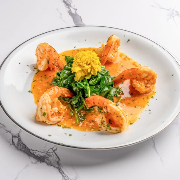 Shrimp scampi with spinach and a yellow edible flower on a white plate with a gold rim.