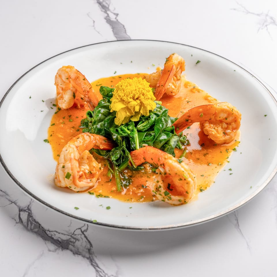 Shrimp scampi with spinach and a yellow edible flower on a white plate with a gold rim.