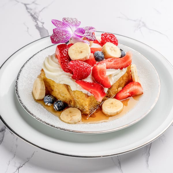 Golden French toast topped with whipped cream, fresh strawberries, blueberries, bananas, and a purple orchid.