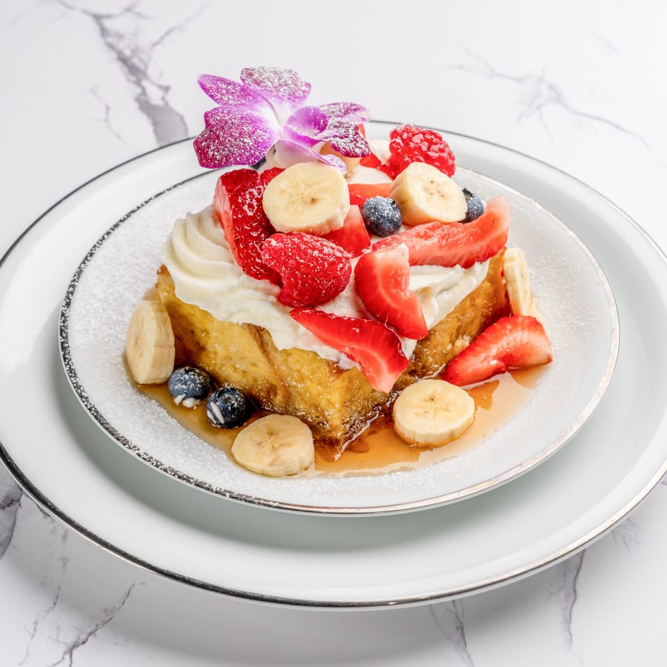 Golden French toast topped with whipped cream, fresh strawberries, blueberries, bananas, and a purple orchid.