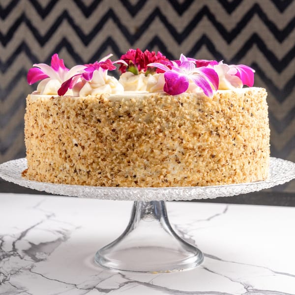 A round cake with nut-covered sides, white frosting, and pink orchid decorations on a glass stand.
