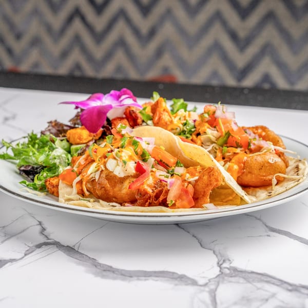 Three fish tacos with salsa, cabbage, and sauce, garnished with a purple orchid and side salad.