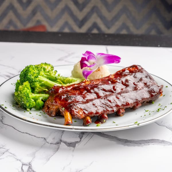 Glazed BBQ ribs with a side of broccoli, mashed potatoes, and a purple orchid garnish.