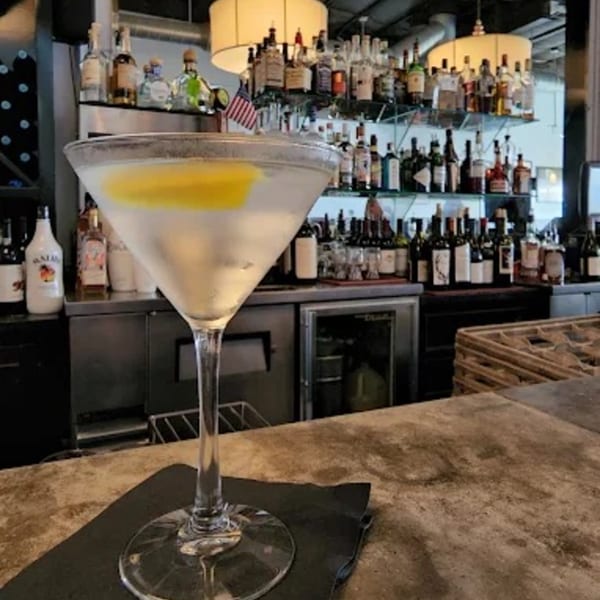 A martini with a lemon twist sits on a bar with bottles and glasses in the background.