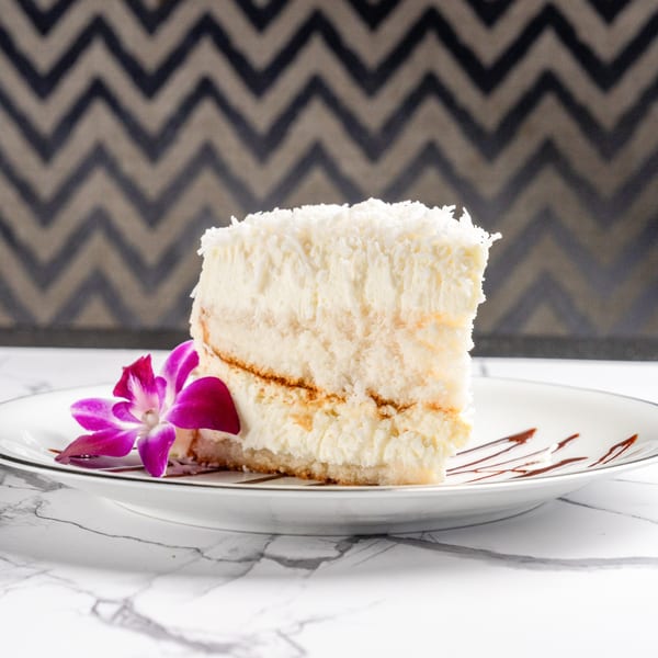 Slice of coconut cake with creamy frosting and a purple orchid on a white plate with chocolate drizzle.