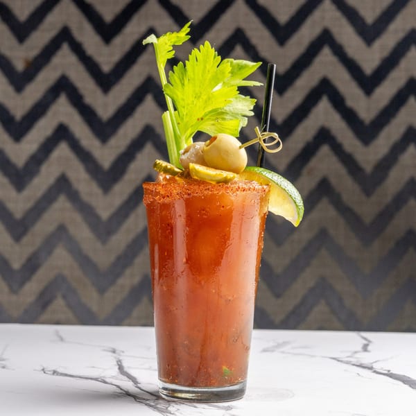 Bloody Mary cocktail garnished with celery, olives, pickles, and a lime wedge on a marble surface.