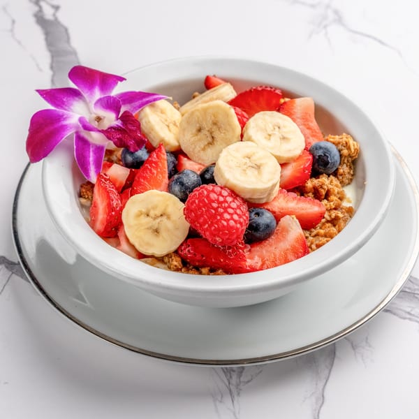 A white bowl of fruit and granola, topped with bananas, strawberries, blueberries, raspberries, and an orchid.