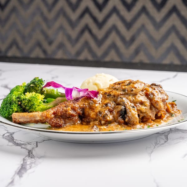 Braised lamb shank with mushroom sauce, mashed potatoes, and broccoli, garnished with a purple orchid.