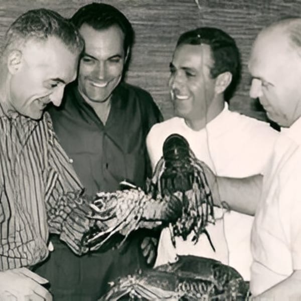 Four smiling men gathered around a table, admiring several large lobsters.