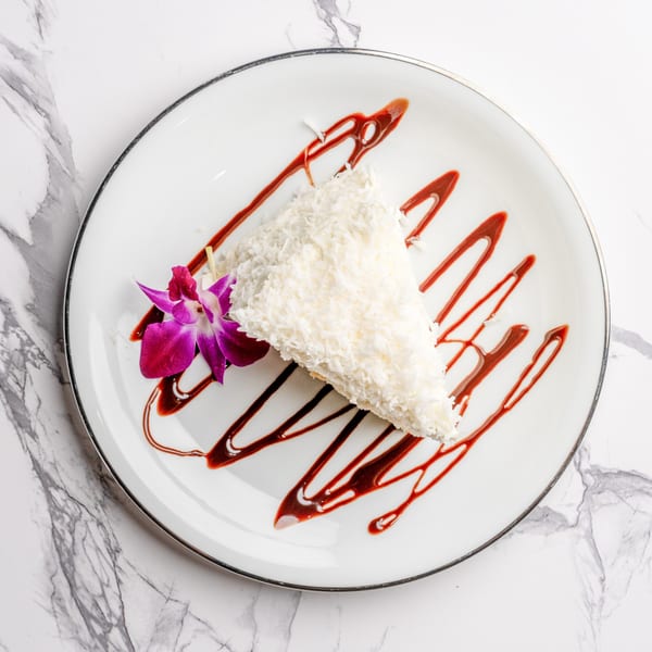 A slice of coconut cake with raspberry drizzle and a purple orchid on a white plate.