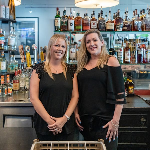 Two smiling women bartenders stand behind a well-stocked bar with liquor bottles and beer taps.