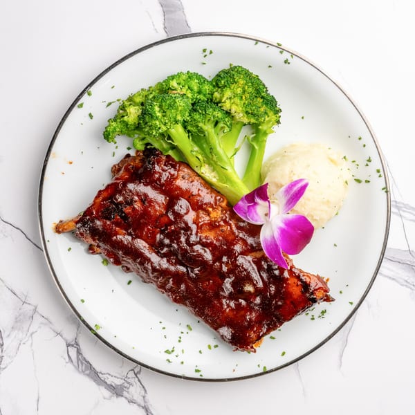 Bistro K's BBQ ribs with mashed potatoes, broccoli, and an orchid garnish on a white plate.