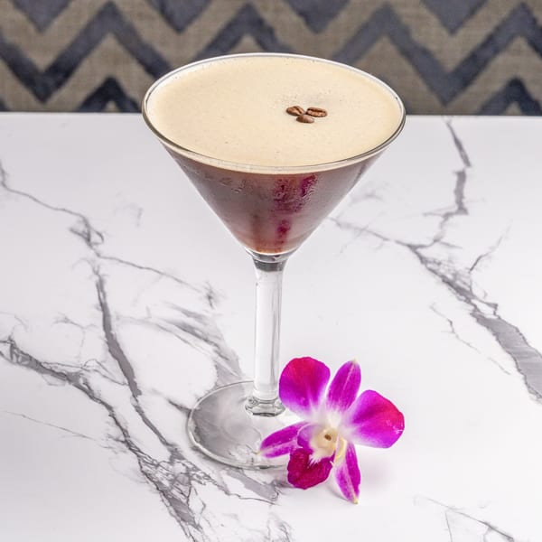Espresso martini in a stemmed glass with coffee beans and a purple orchid on a marble table.