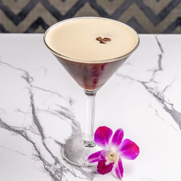 Espresso martini with coffee beans and a purple orchid on a white marble table.