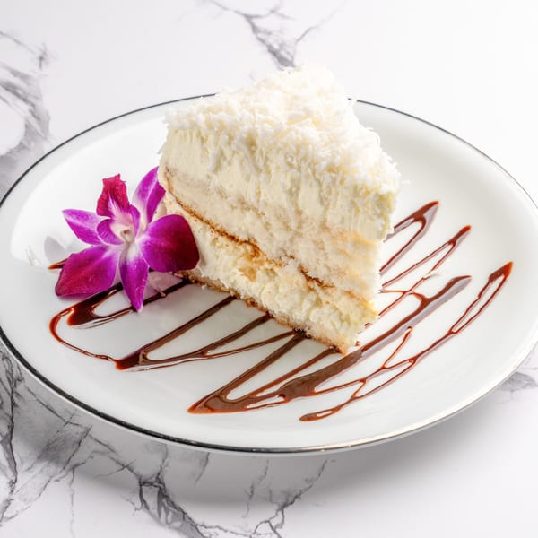 A slice of coconut cake with chocolate drizzle and a purple orchid on a white plate.