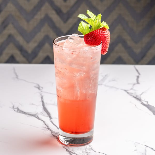 A tall glass of strawberry Italian soda with ice, garnished with a strawberry and mint.