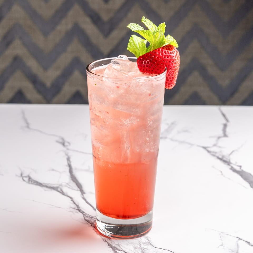A tall glass of strawberry Italian soda with ice, garnished with a strawberry and mint.