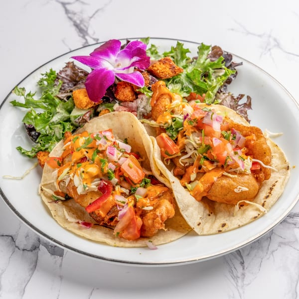 Two fish tacos with sauce, pico de gallo, and a side salad with a purple orchid garnish.