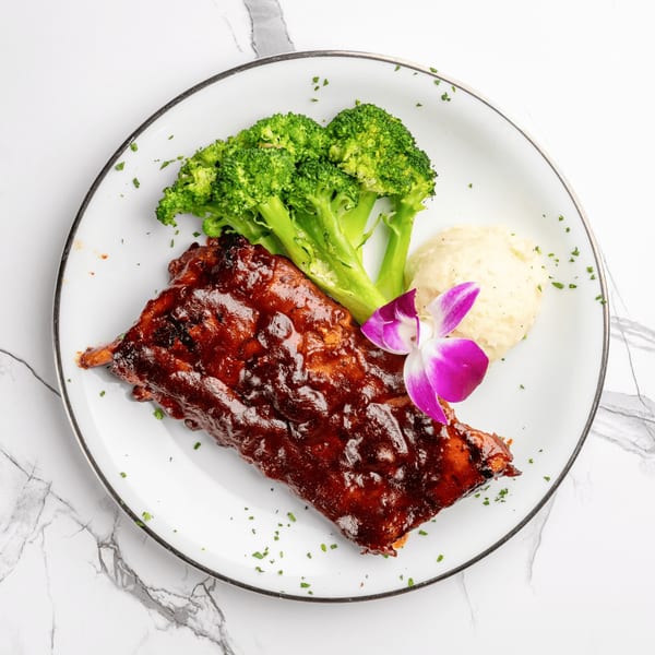 Ribs with barbecue sauce, mashed potatoes, broccoli, and a purple orchid on a white plate.